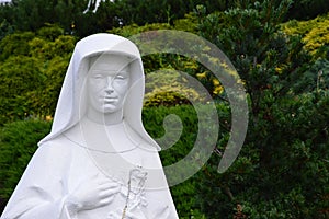 Detail of sculpture of polish Saint Faustyna Kowalska holding cross and rosary, decorative shrubs in background.