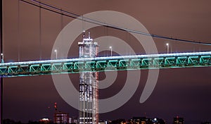 Detail of suspension bridge at night with high rise construction in the background..