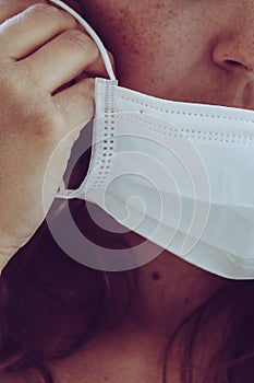 Detail of unrecognizable woman face wearing white medical face mask. Partly blurred, selective focus, light grain. Redhead girl