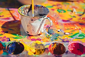 Detail view of messy painting colours and painter`s cup with paintbrush on colorful canvas