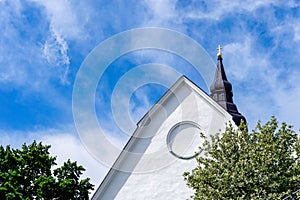 Detail view of a whitewashed church building with steeple under a blue sky with white clouds and green trees below