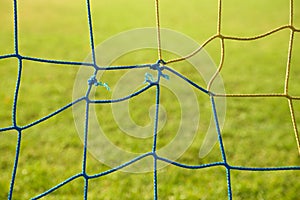 Detail of yellow blue crossed soccer nets, soccer football in goal net with poor grass on playground in background.