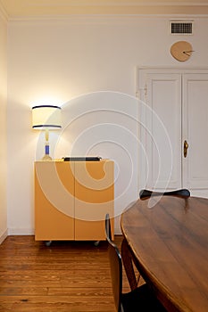 Detail yellow sideboard in kitchen with lighted lamp and wooden table with black chairs. Antique apartment interior with modern