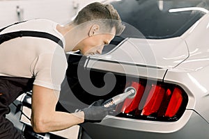 Detailing and polishing of car tail light on car. Young professional concentrated Caucasian male worker with orbital