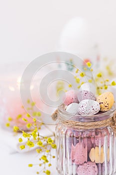 Details of festive Easter table setting, chocolate candy Easter eggs in pastel colors in crystal jar, candle, flowers