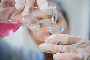 Details: Gloved hands of laboratory personnel pouring pink liquid chemical substance from a glass flask through a funnel