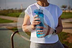 Details on the hands of a sportswoman holding a bottle of water, renewing qua balance after a heavy workout outdoors