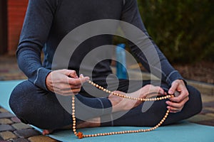 Details: hands of a yogi sitting on a fitness mat, meditating with rosary beads while practicing yoga outdoors.