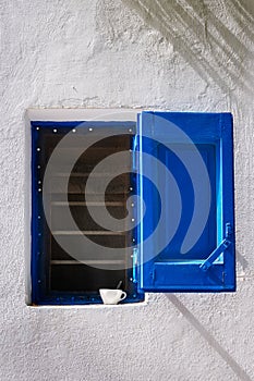 Details of traditional Greek island house. Whitewashed wall, small window, blue storm shutter open, coffee cup on window