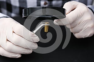 Detective exploring bullet shell with magnifying glass on black background, closeup