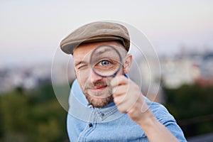 Detective male in peaked cap making big eye using loupe