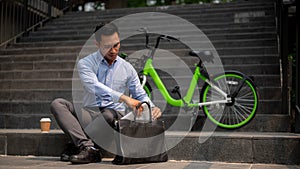 A determined Asian businessman sits on steps outdoors, organizing his laptop in his briefcase bag