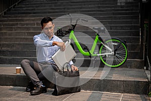 A determined Asian businessman sits on steps outdoors, organizing his laptop in his briefcase bag