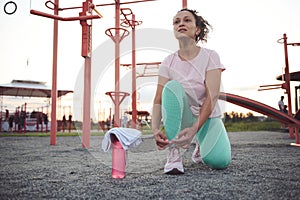 Determined middle-aged sportswoman tying shoelaces, on an outdoor sports fields against gym machines at sunset