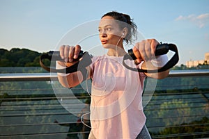 Determined sportswoman with athletic physique doing exercises on arms with suspension straps while performing a bodyweight