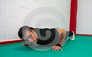 Determined strong young man doing push-ups and looking at camera. He is dressed in an orange shirt and black jumpsuit