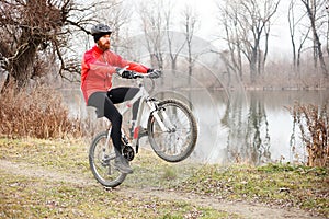 Determined young bearded man doing a wheelie on a mountain bike by the river