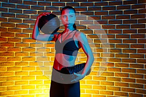 Determined young brunette woman posing with medicine ball and foam fitness roller in gym in neon lights.