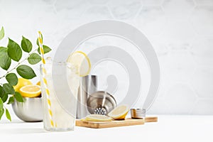 Detox fresh lemon juice cooking - lemonade of ripe lemons with ice in glass with silver bowl, shaker on white table, marble tile.