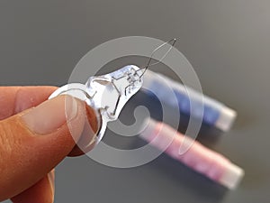 Device for drawing the thread into the eye of the needle. Bobbins with blue and pink threads in the background. A woman holds a