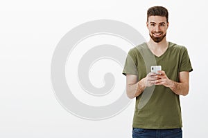 Devious and excited good-looking mature bearded male in olive t-shirt holding smartphone and looking pleased at camera