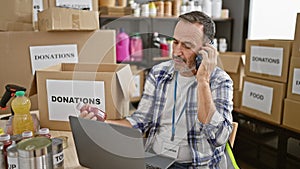 Devoted middle age man with grey hair, earnestly talking on smartphone while managing donations via laptop at bustling charity