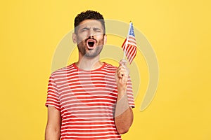 Devoted patriotic man in striped t-shirt holding in hand flag of united states of america, singing hymn celebrating independence