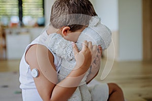 Diabetic boy with a continuous glucose monitor sitting at home, hugging his stuffed teddy bear.