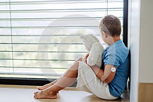 Diabetic boy with a continuous glucose monitor sitting by the window, holding his stuffed teddy bear and looking outside