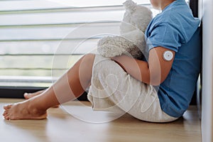 Diabetic boy with a continuous glucose monitor sitting by the window, holding his stuffed teddy bear and looking outside