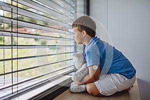 Diabetic boy with a continuous glucose monitor sitting by the window, holding his stuffed teddy bear and looking outside