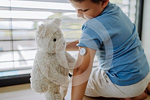 Diabetic boy with a continuous glucose monitor sitting by the window, showing his stuffed teddy bear sensor on his arm.