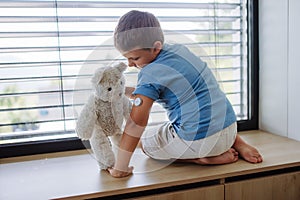 Diabetic boy with a continuous glucose monitor sitting by the window, showing his stuffed teddy bear sensor on his arm.