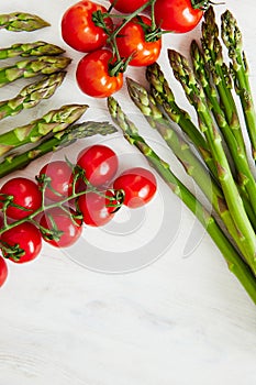 Diagonal composition of asparagus stems and branches of cherry tomatoes on a off-white rustic table.