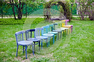 a diagonal row of rainbow wooden chairs on a green lawn in the park, preparing for a family party on a weekend in the