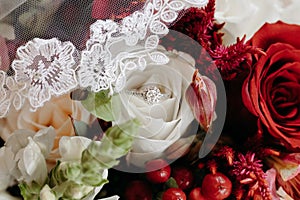 Diamond engagement ring on white rose with bride& x27;s veil and red roses. Bridal bouquet with wedding ring and red berries