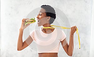 Diet breakdown. Black lady trying to eat unhealthy burger, pulling herself away by measuring tape, grey background