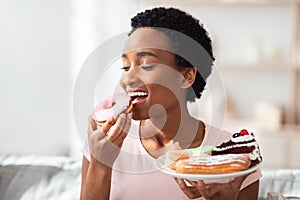 Diet breakdown, cheat meal and imbalanced nutrition. Young black woman with plate of sweets eating donut at home