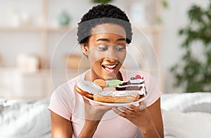 Diet breakdown and unhealthy nutrition concept. Young African American lady holding plate of sweet pastries at home