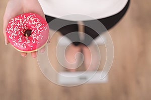 Diet Concept. Young Woman Measuring Body Weight On Weighing Scale While Holding Glazed Donut With Sprinkles. Sweets Are Unhealthy