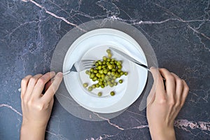 Diet. A hand with a fork and a table knife eats canned peas from a white plate, top view