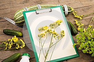 marrow squash, measure tape, blank clipboard, bottle of water, yellow flowers and cucumbers on brown wooden table