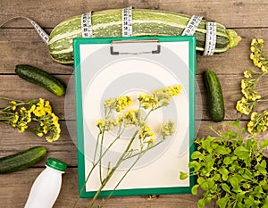 marrow squash, measure tape, blank clipboard, bottle of water, yellow flowers and cucumbers on brown wooden table