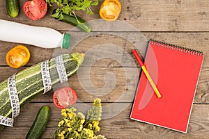 marrow squash, measure tape, blank red notepad, bottle of water, flowers, tomatoes and cucumbers on brown wooden table