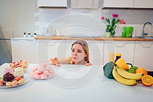 Diet struggle. Young sad woman in blue T-shirt choosing between fresh fruit vegetables or sweets in the kitchen. Choice