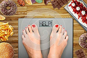 Diet temptation or hard to lose weight concept with woman weighing on bathroom scale with many sweets and fast food around