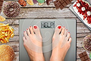 Diet temptation or hard to lose weight concept with woman weighing on bathroom scale with many sweets and fast food around