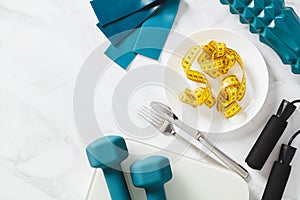Diet, weight loss concept. Flat lay plate with centimetre tape, cutlery, dumbbells, scales and fitness equipment on white marble photo