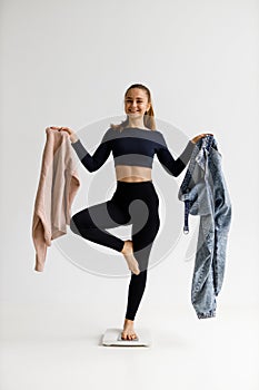 Diet and weight loss. A young woman holds oversized denim pants and a jacket in her hands and stands on the scales