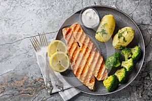 Dietary grilled salmon with boiled potatoes, broccoli, lemon, herbs and cream sauce close-up in a plate. Horizontal top view